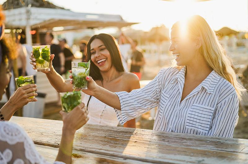 Amiche brindano sorridenti al tramonto sulla spiaggia, atmosfera di gioia e relax estivo.